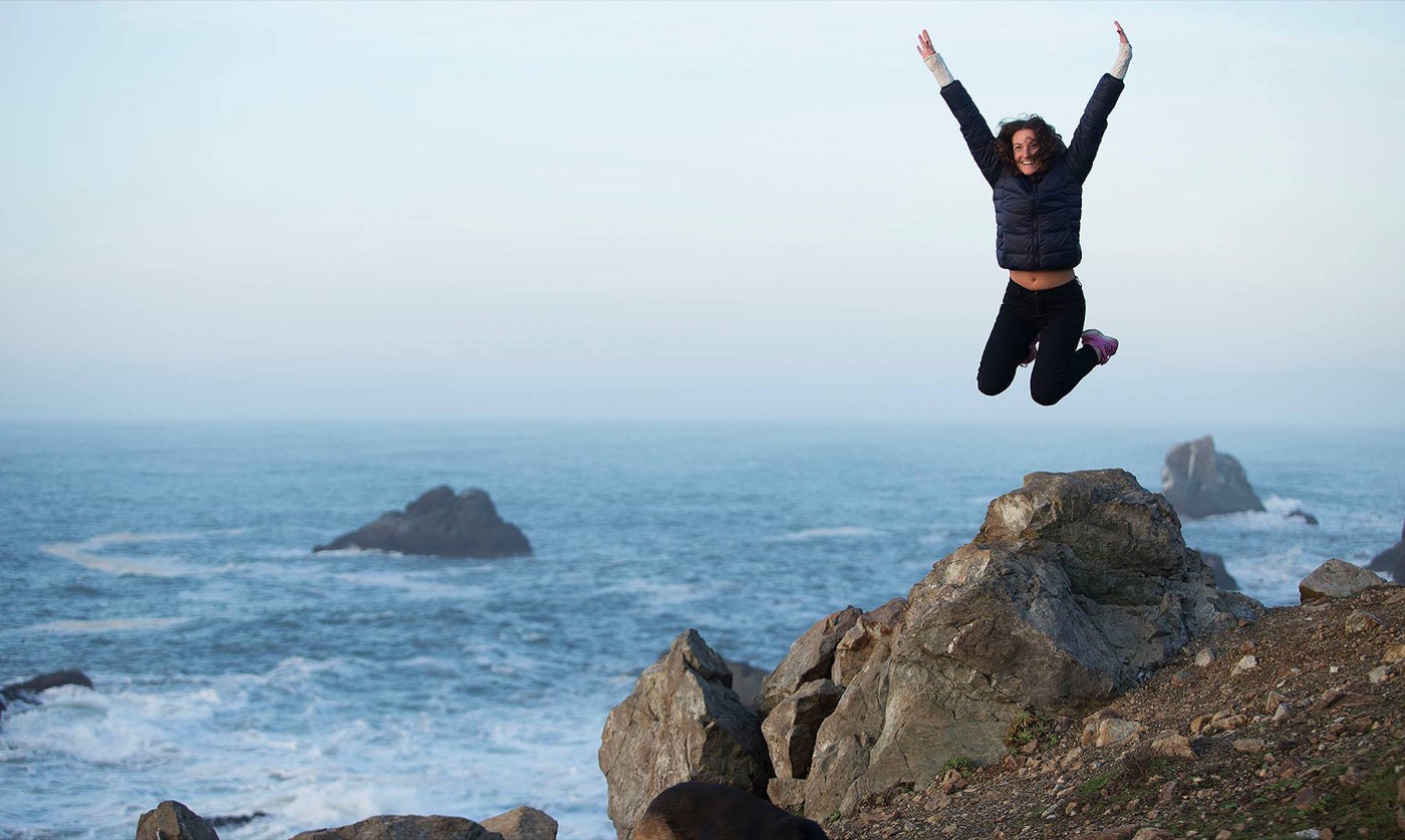 A woman jumping for joy on a rocky point above the Pacific Ocean