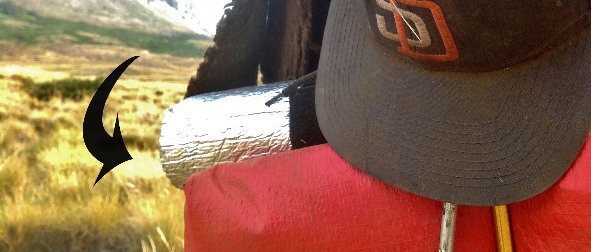 A rolled red tarp strapped to a backpack next to a San Diego Padres cap, grassland behind