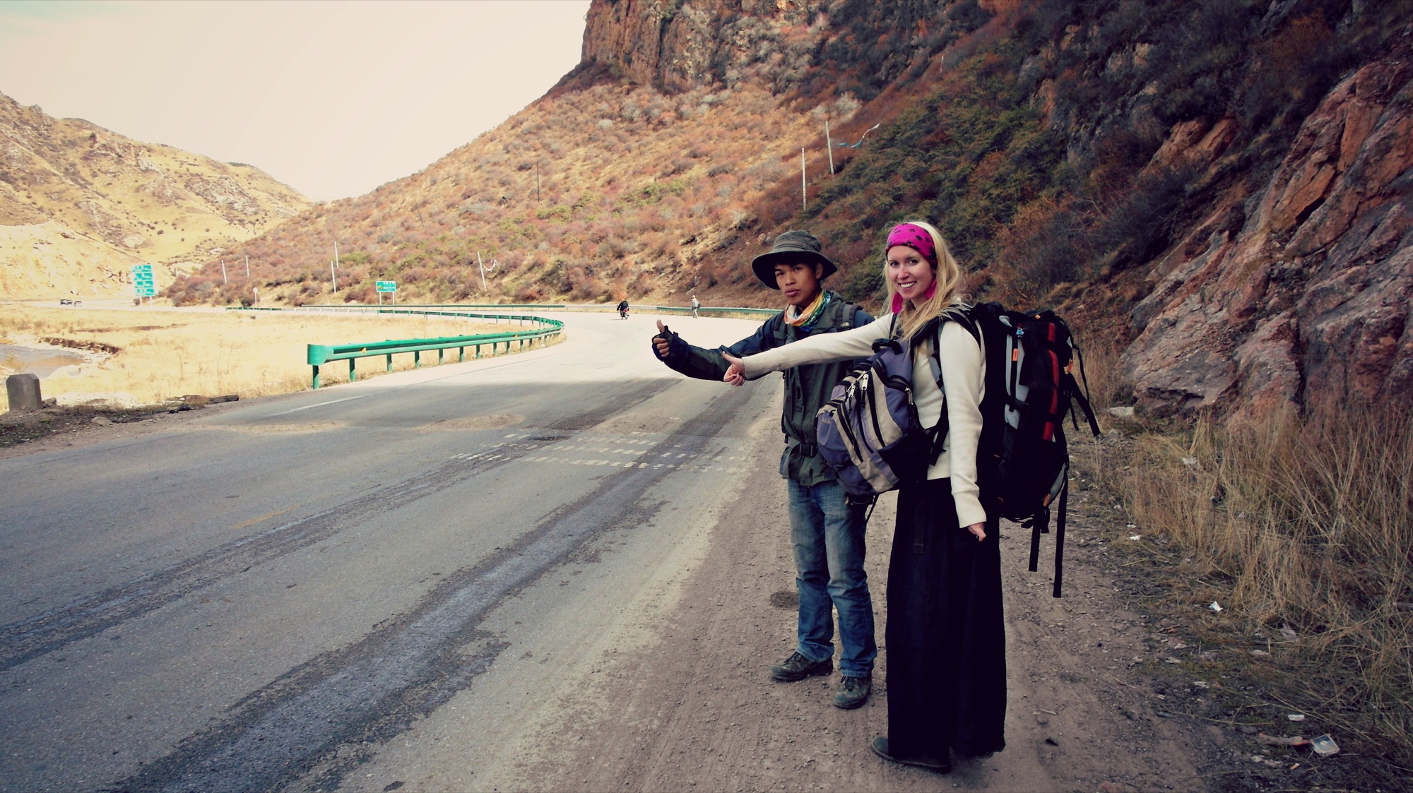 Two backpackers hitchhiking on the shoulder of a mountain road