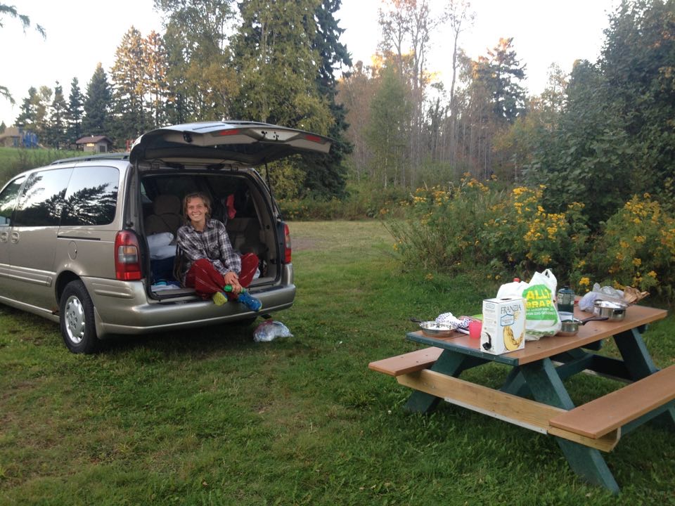 Olivia sitting in the open back of a minivan at a campsite, next to a picnic table